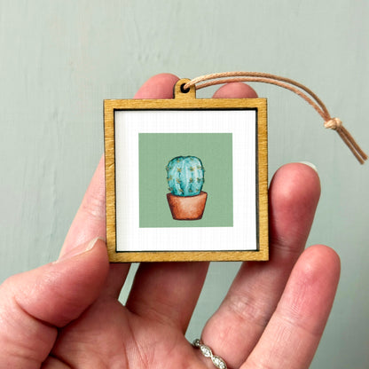 A hand holding a small wooden frame containing a framed illustration of a cactus plant.