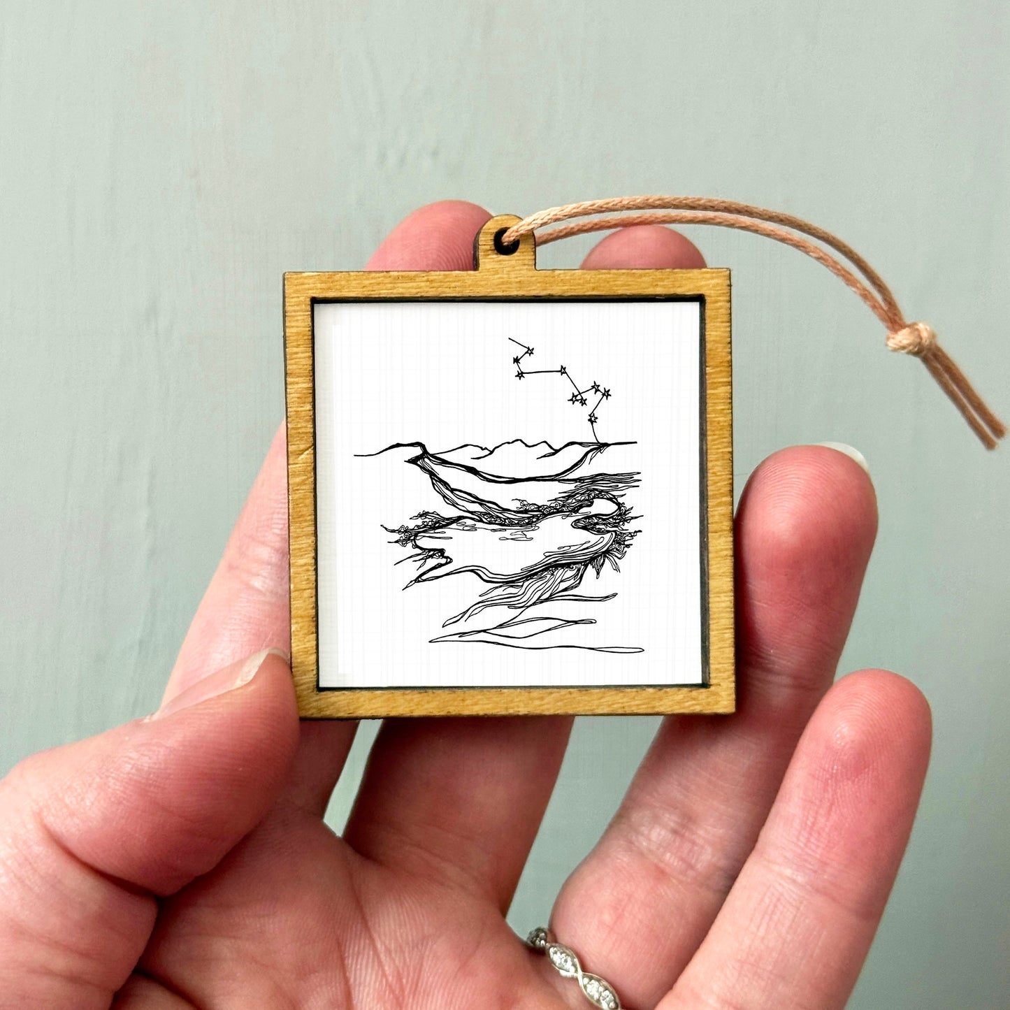A hand holding a small wooden frame containing a black and white line drawing of a landscape with mountains and a body of water.
