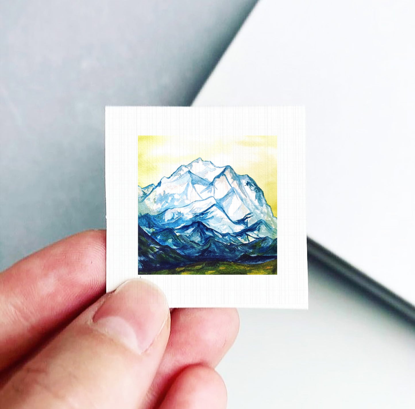 A hand is holding a small square piece of paper with a painting of a mountain landscape on it.