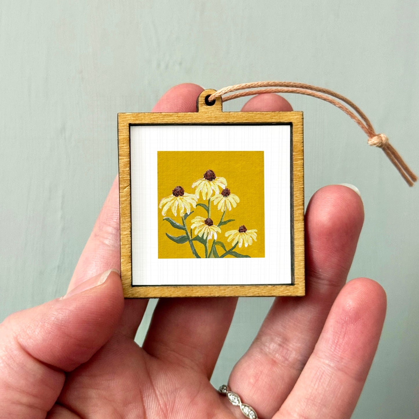 A hand holding a small wooden frame containing a painting of yellow flowers.