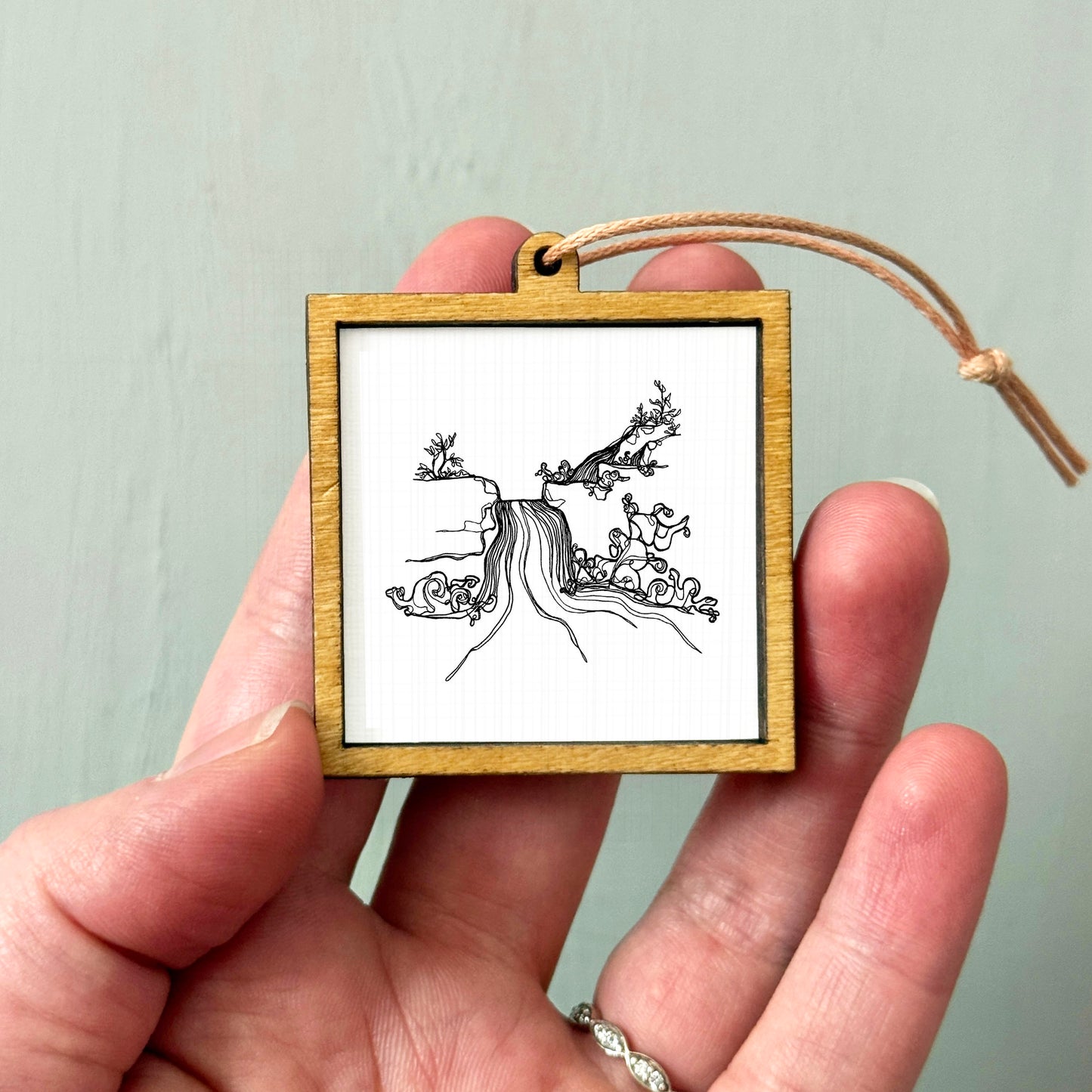 A hand holding a small wooden frame containing a black and white illustration of a waterfall.