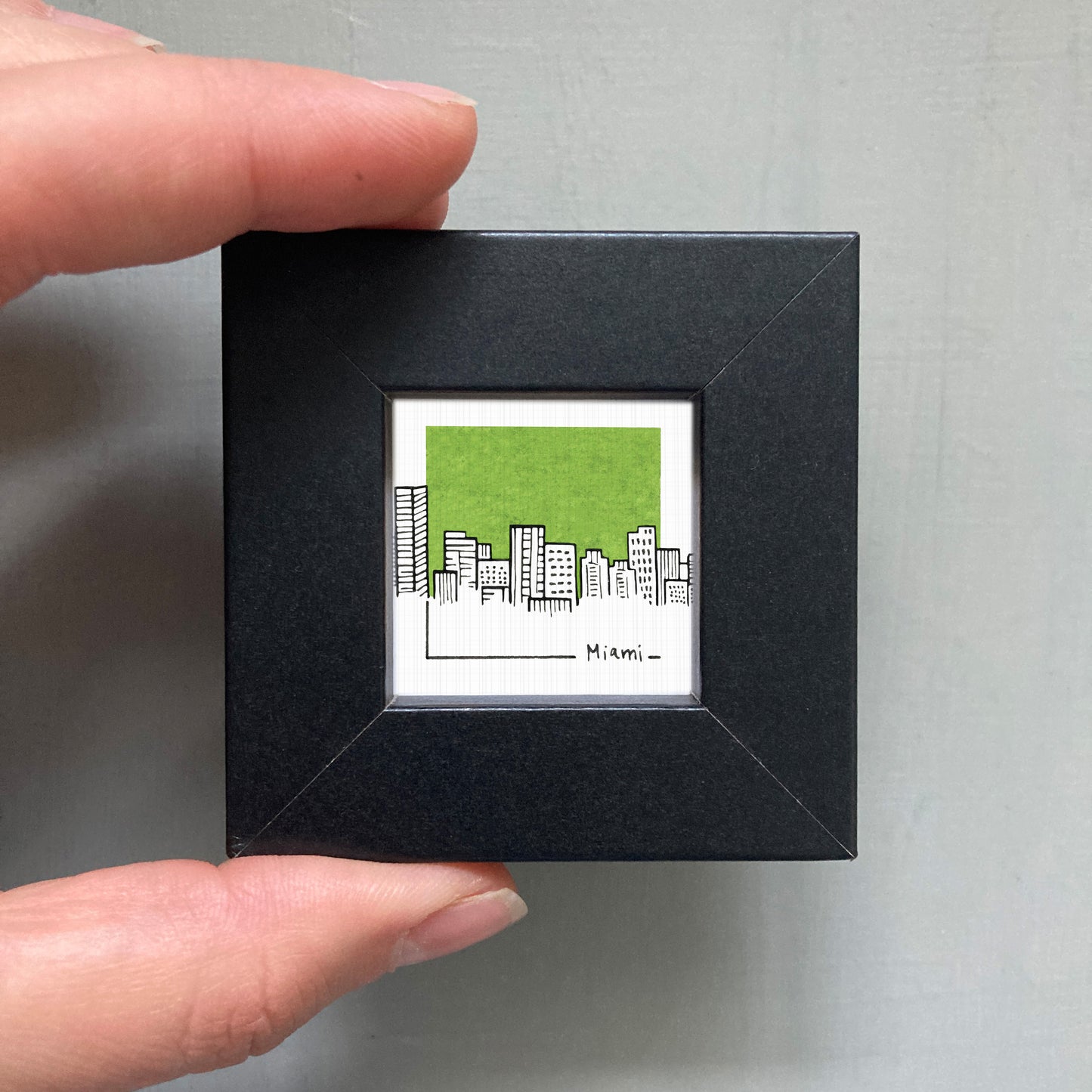 A person's hand holding a small black frame containing a miniature illustration of a city skyline with green buildings.