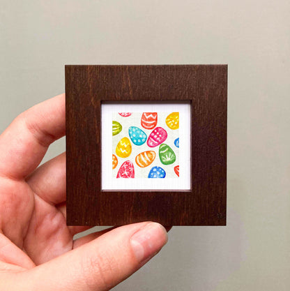 A hand is holding a small square frame containing a colorful arrangement of Easter eggs.