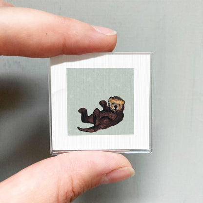 A person's hand is holding a small square picture of a brown otter.