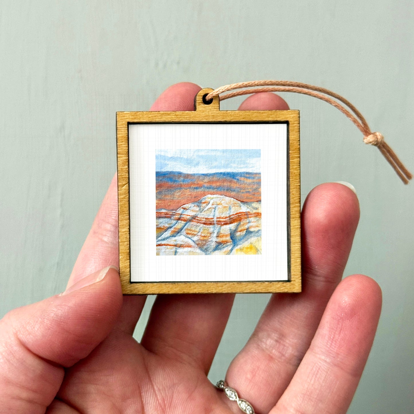 A hand holding a small wooden frame containing a watercolor painting of a mountain landscape.