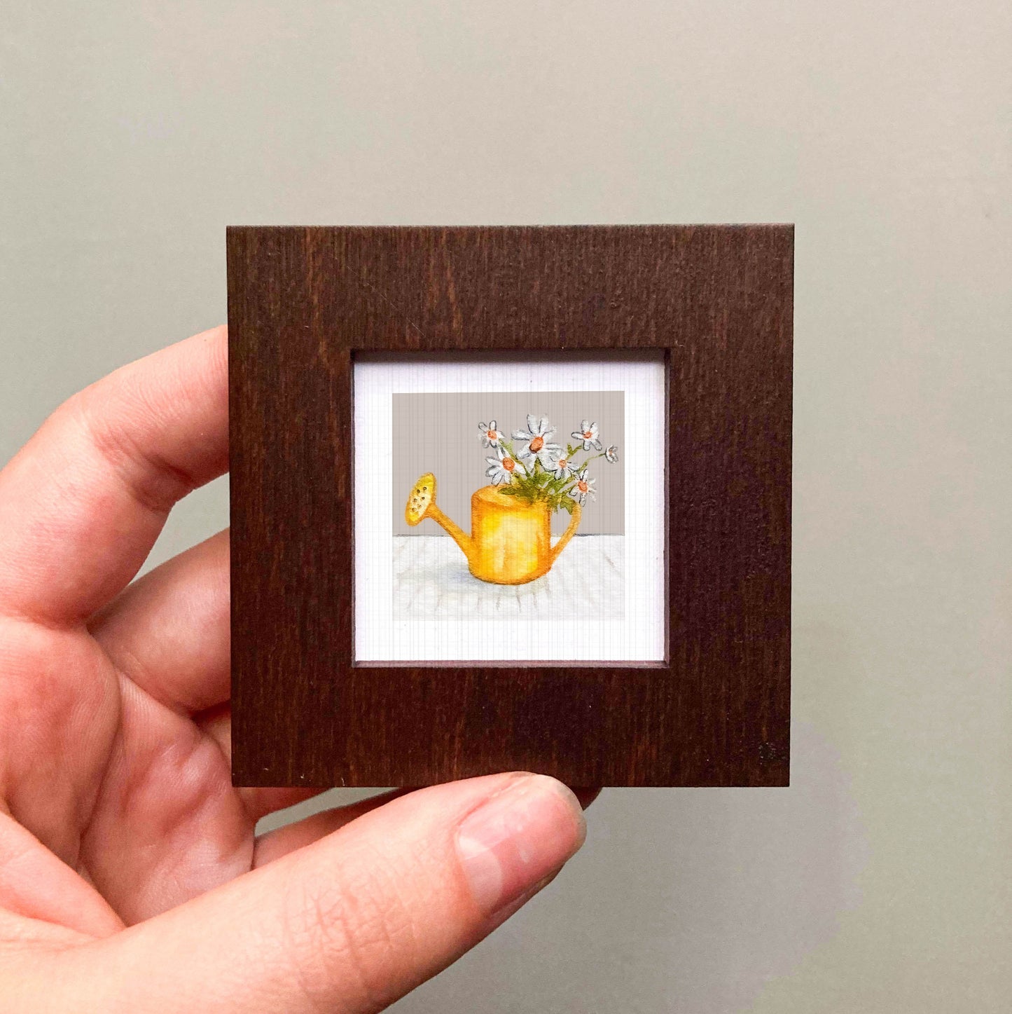 A hand is holding a small, square-shaped picture frame containing a painting of a yellow watering can with flowers.