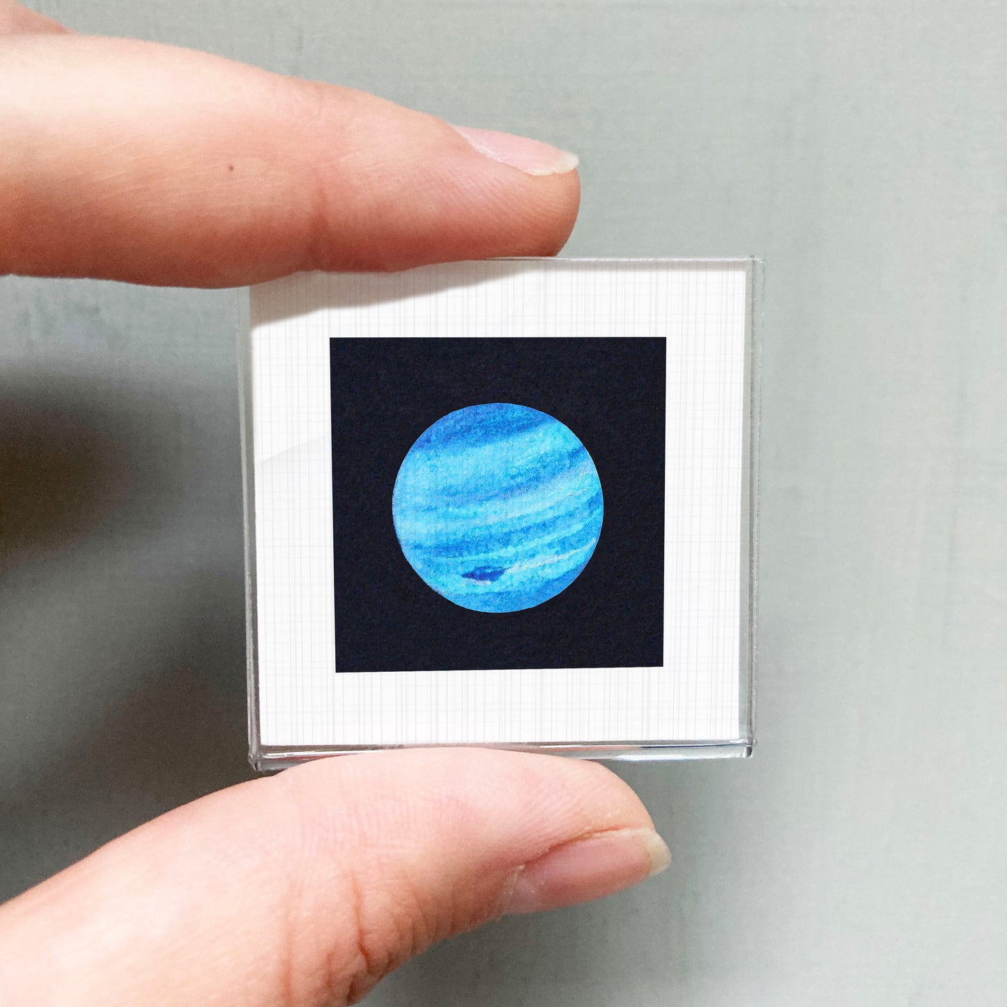 A person's hand is holding a small square frame containing a picture of a blue planet.