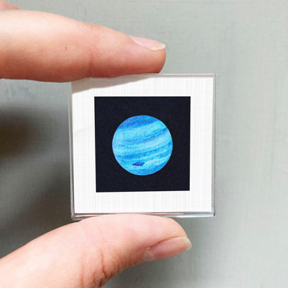 A person's hand is holding a small square frame containing a picture of a blue planet.