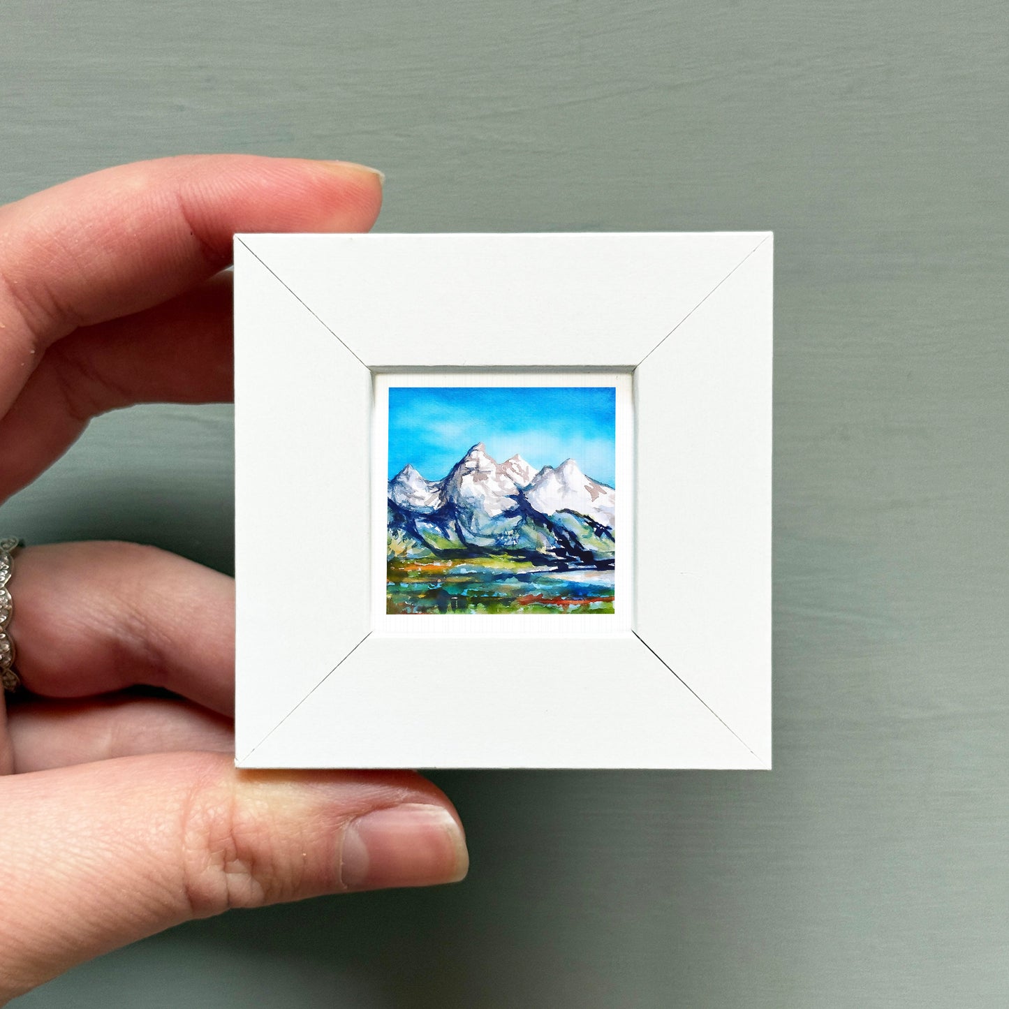 A hand is holding a small white picture frame containing a landscape painting of mountains.