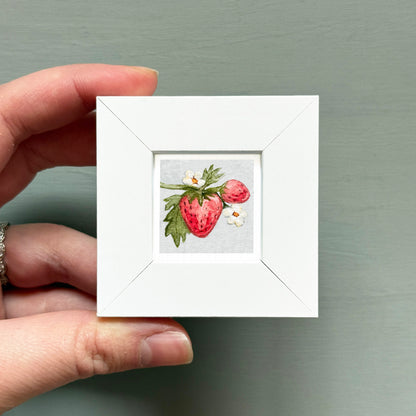 A hand is holding a small white square frame containing a painting of a strawberry with white flowers.