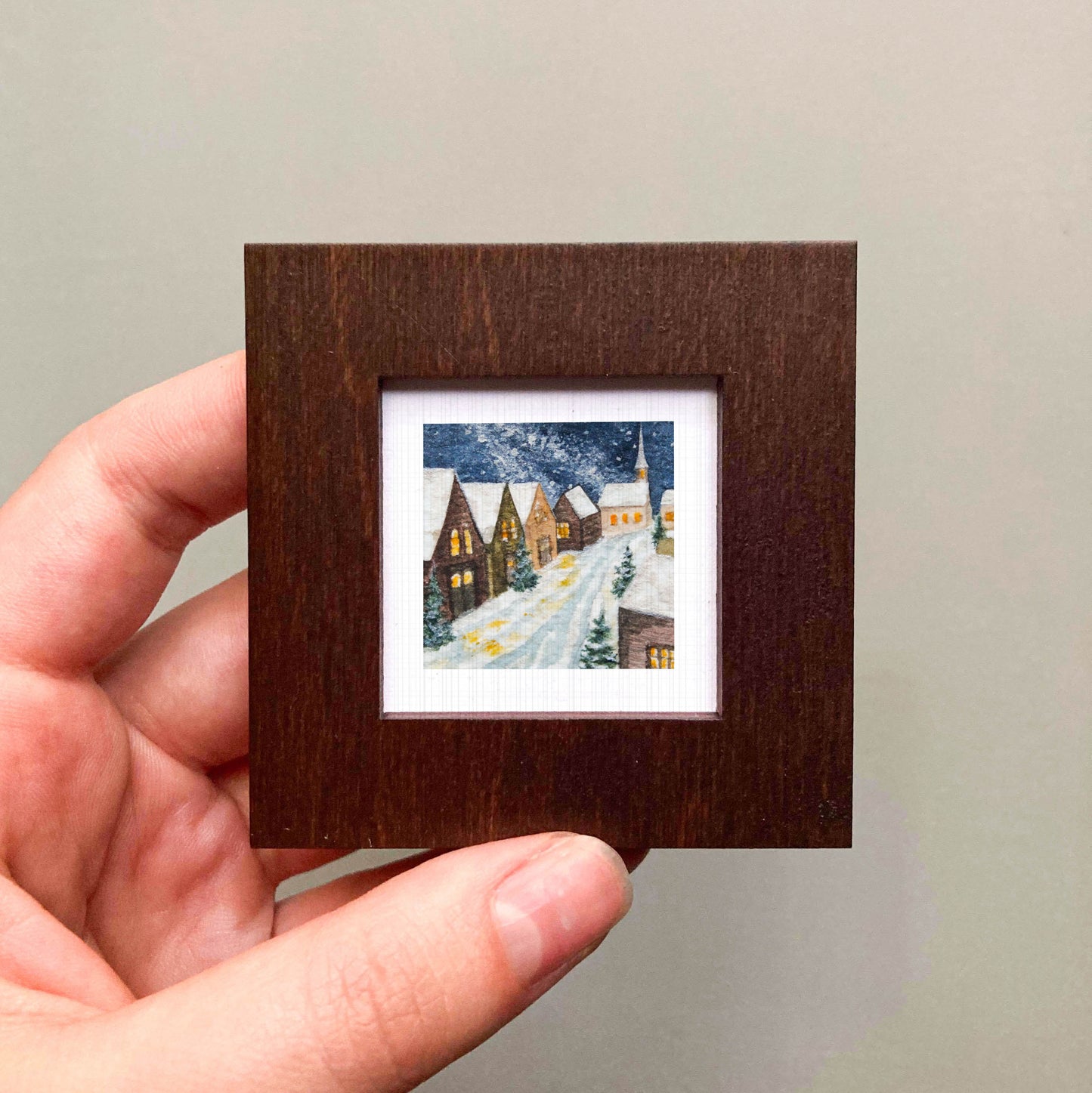 A hand is holding a small, square-shaped wooden frame containing a miniature painting of a snowy village scene.