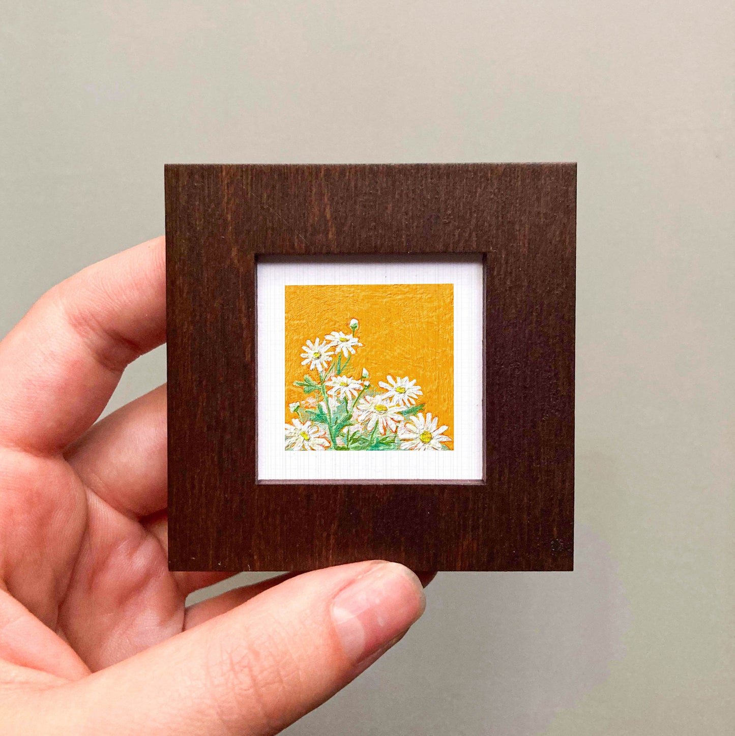 A hand is holding a small wooden frame containing a framed painting of white flowers against a yellow background.