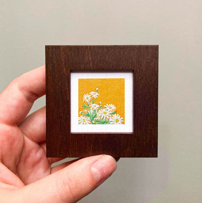 A hand is holding a small wooden frame containing a framed painting of white flowers against a yellow background.
