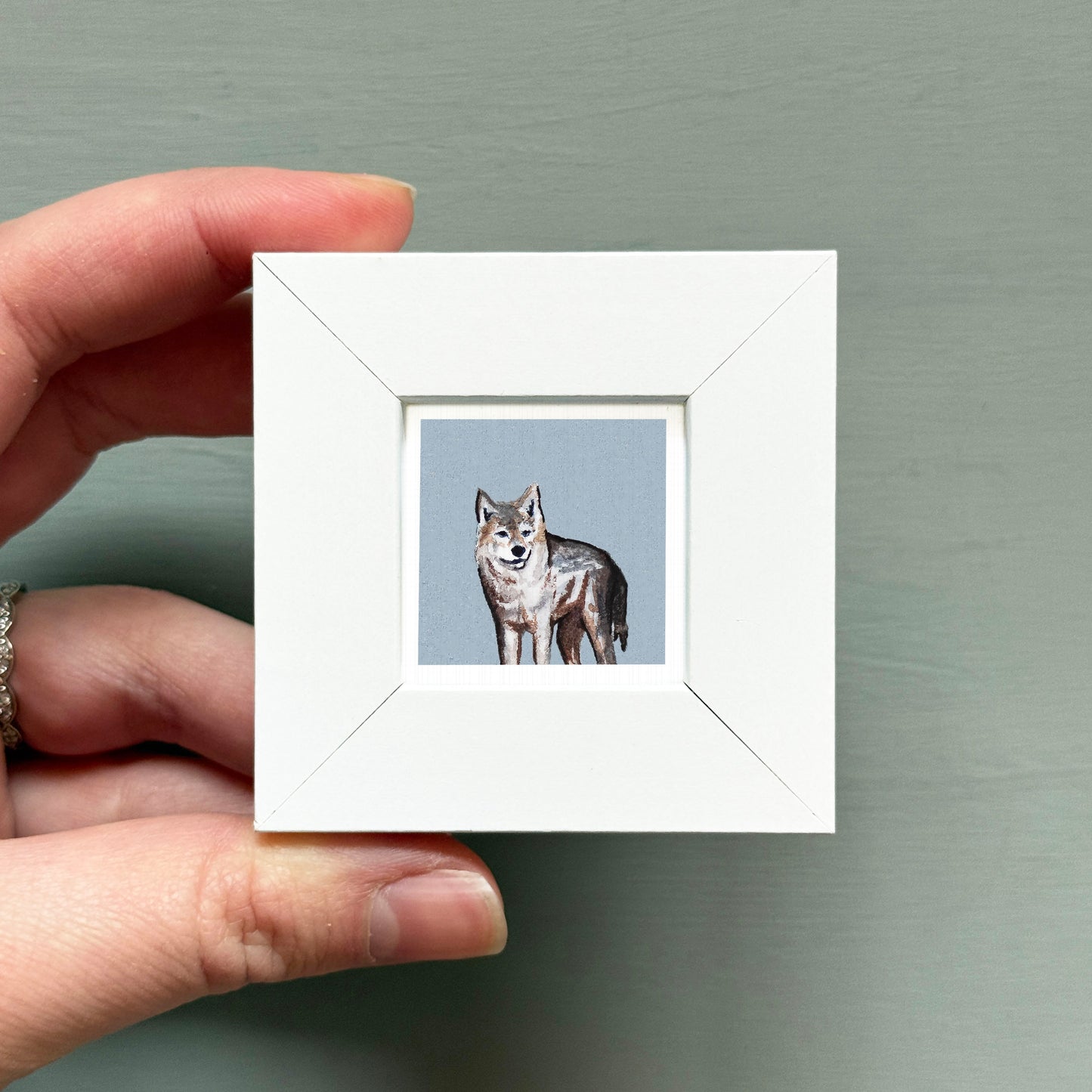 A hand is holding a small white square frame containing a painting of a wolf.