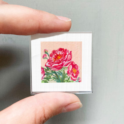 A person's hand holding a small square picture of a vibrant pink flower.
