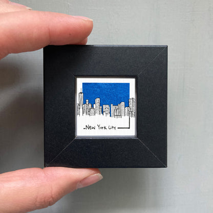 A hand is holding a small square frame containing a drawing of a city skyline with a blue sky.
