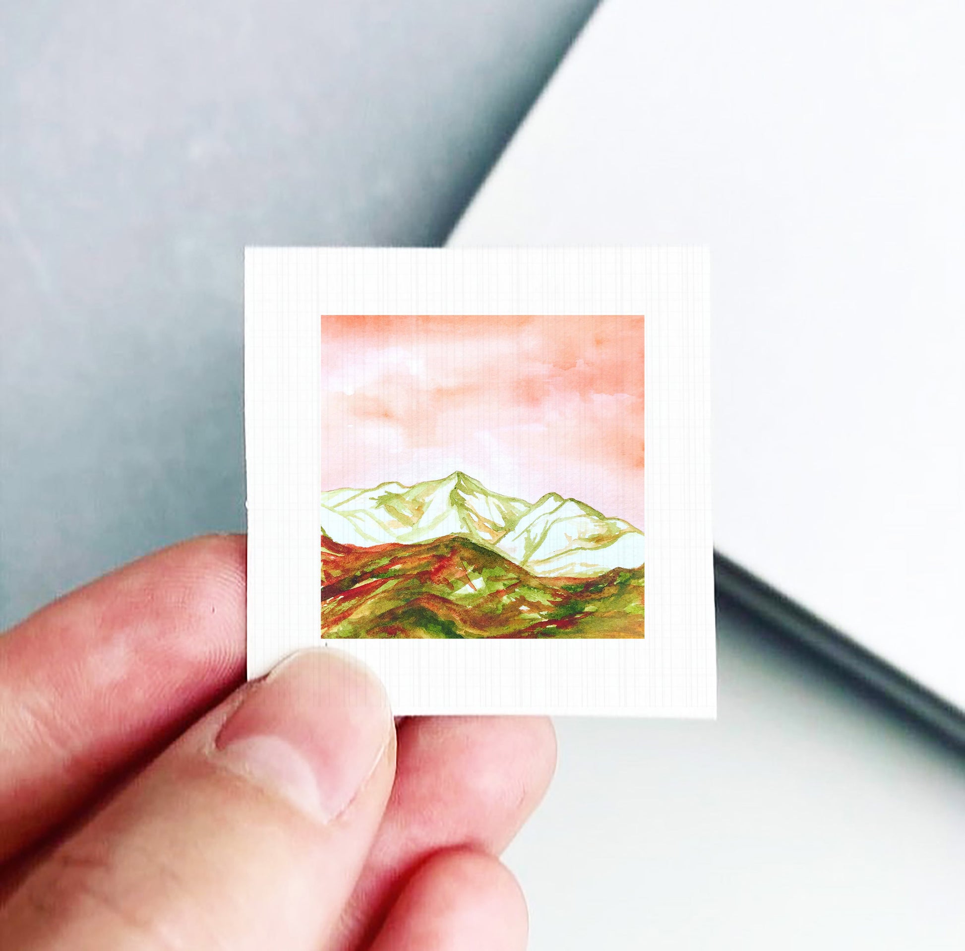 A hand is holding a small square card with a painting of a mountain landscape on it.