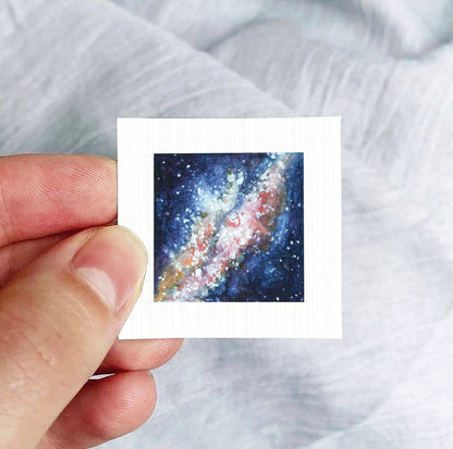 A hand is holding a small square-shaped piece of paper with a painting of a galaxy on it.