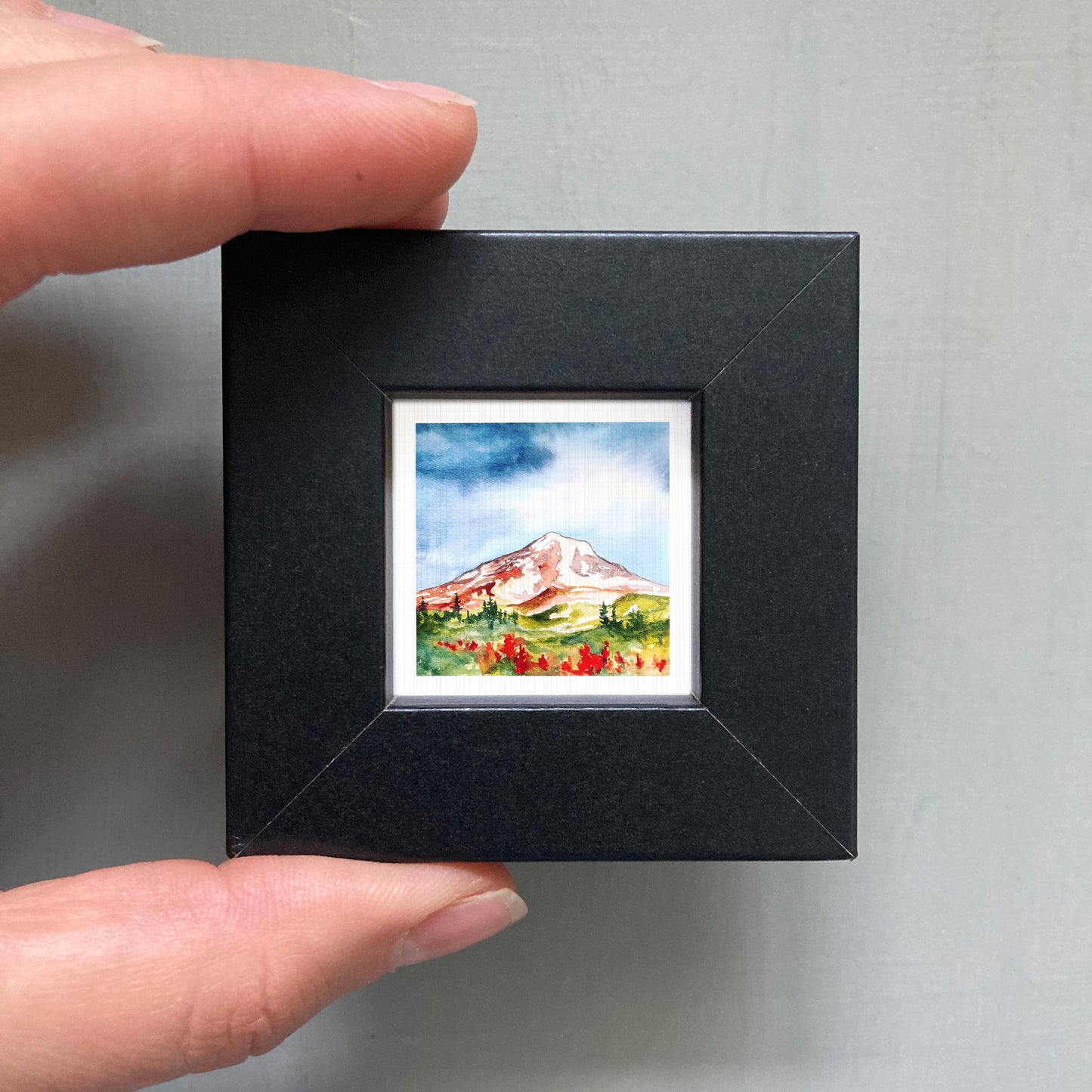 A person's hand holding a small black frame containing a watercolor painting of a mountain landscape.