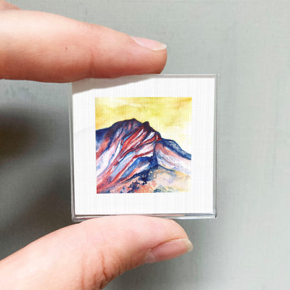A person's hand is holding a small square picture of a mountain landscape with vibrant colors.