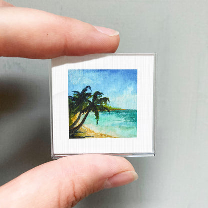 A person's hand holding a small square picture of a tropical beach scene with palm trees and a blue sky.