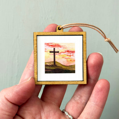 A person's hand holding a small wooden frame containing a framed painting of a cross against a sunset landscape.