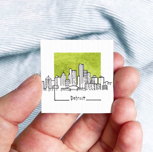A hand holding a small square-shaped paper with a drawing of a city skyline and the word "Detroit" written on it.