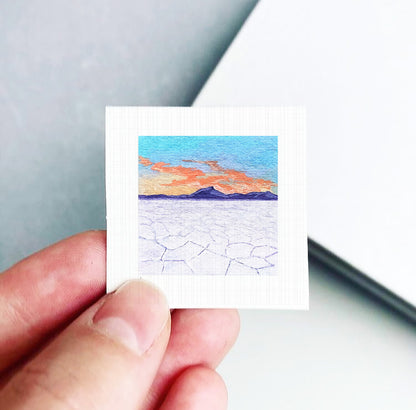 A hand is holding a small square picture depicting a landscape with mountains and a colorful sky.