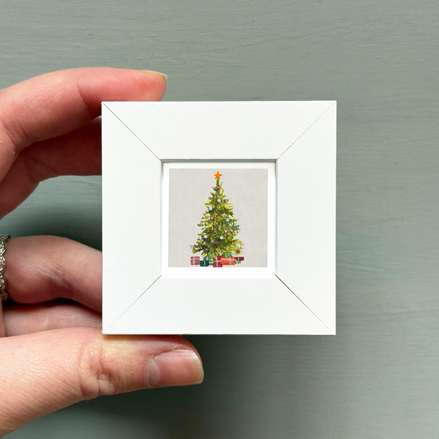 A hand is holding a small white square frame containing a miniature Christmas tree with presents underneath.