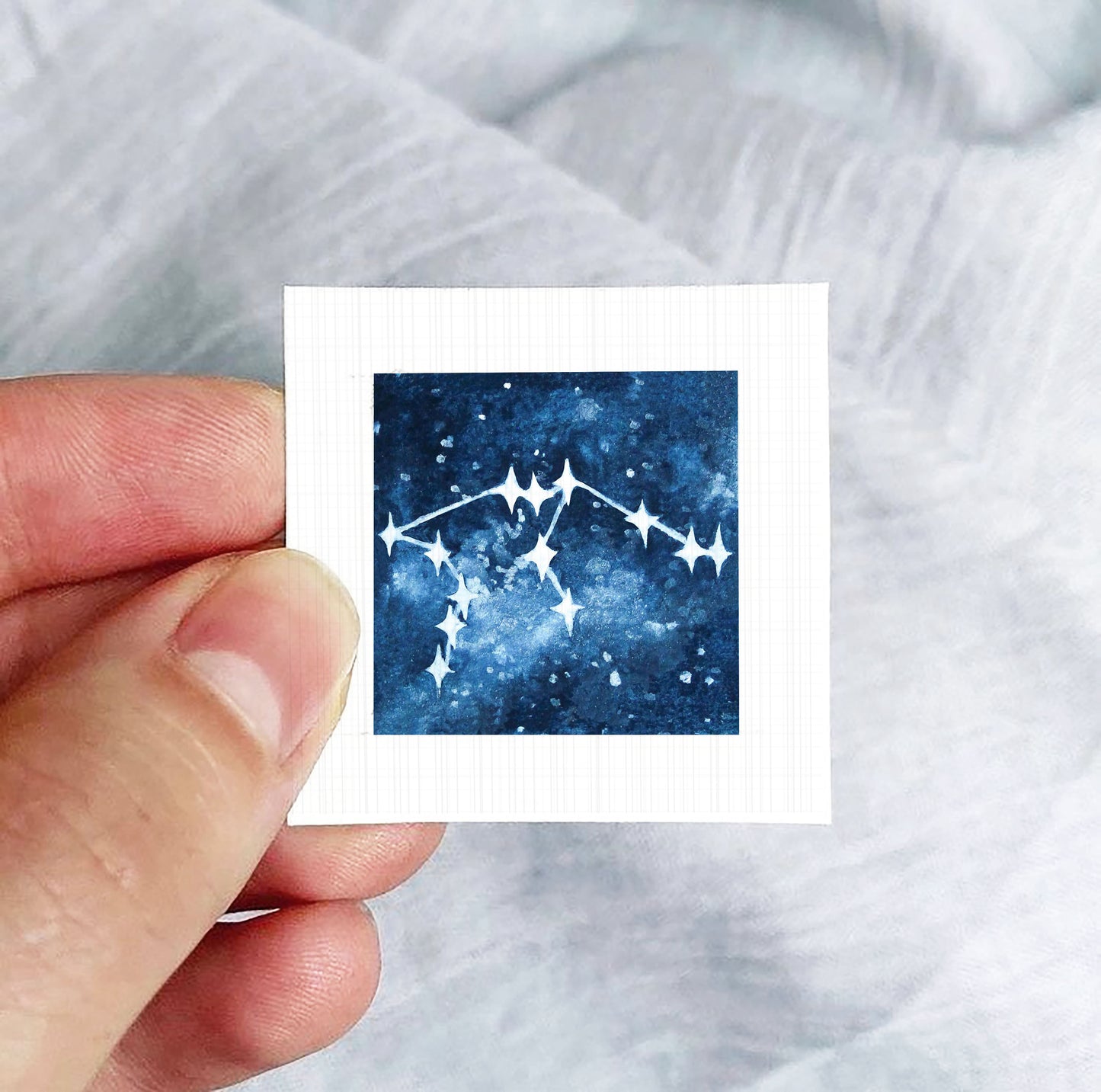 A hand is holding a small square-shaped paper with a depiction of a starry night sky on it.