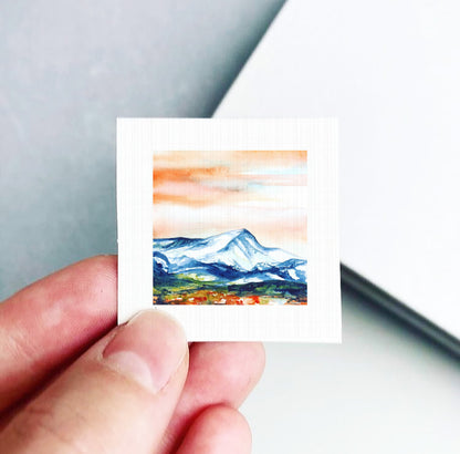 A hand is holding a small square artwork depicting a mountain landscape with a colorful sky.