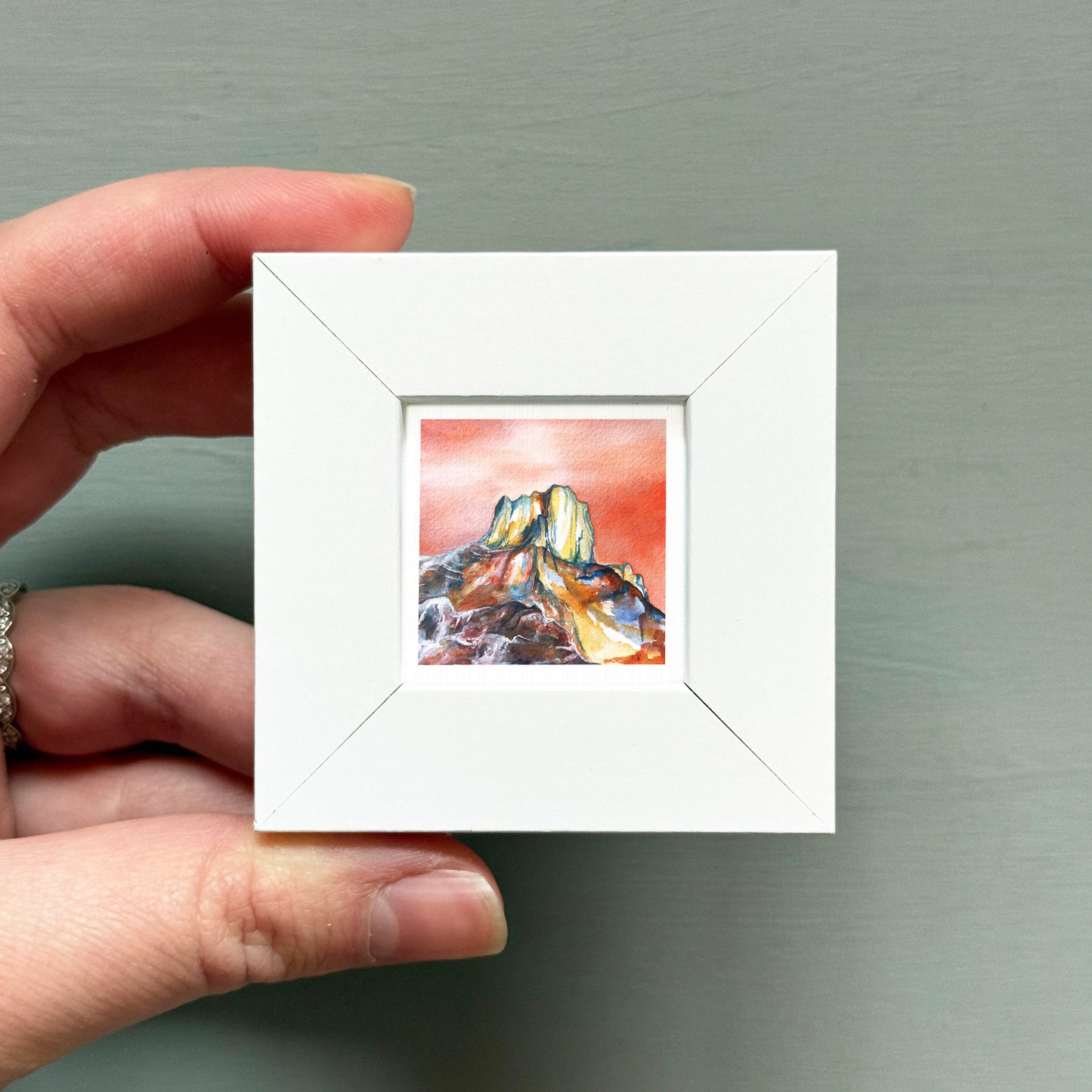 A hand is holding a small white square frame containing a watercolor painting of a mountain landscape.