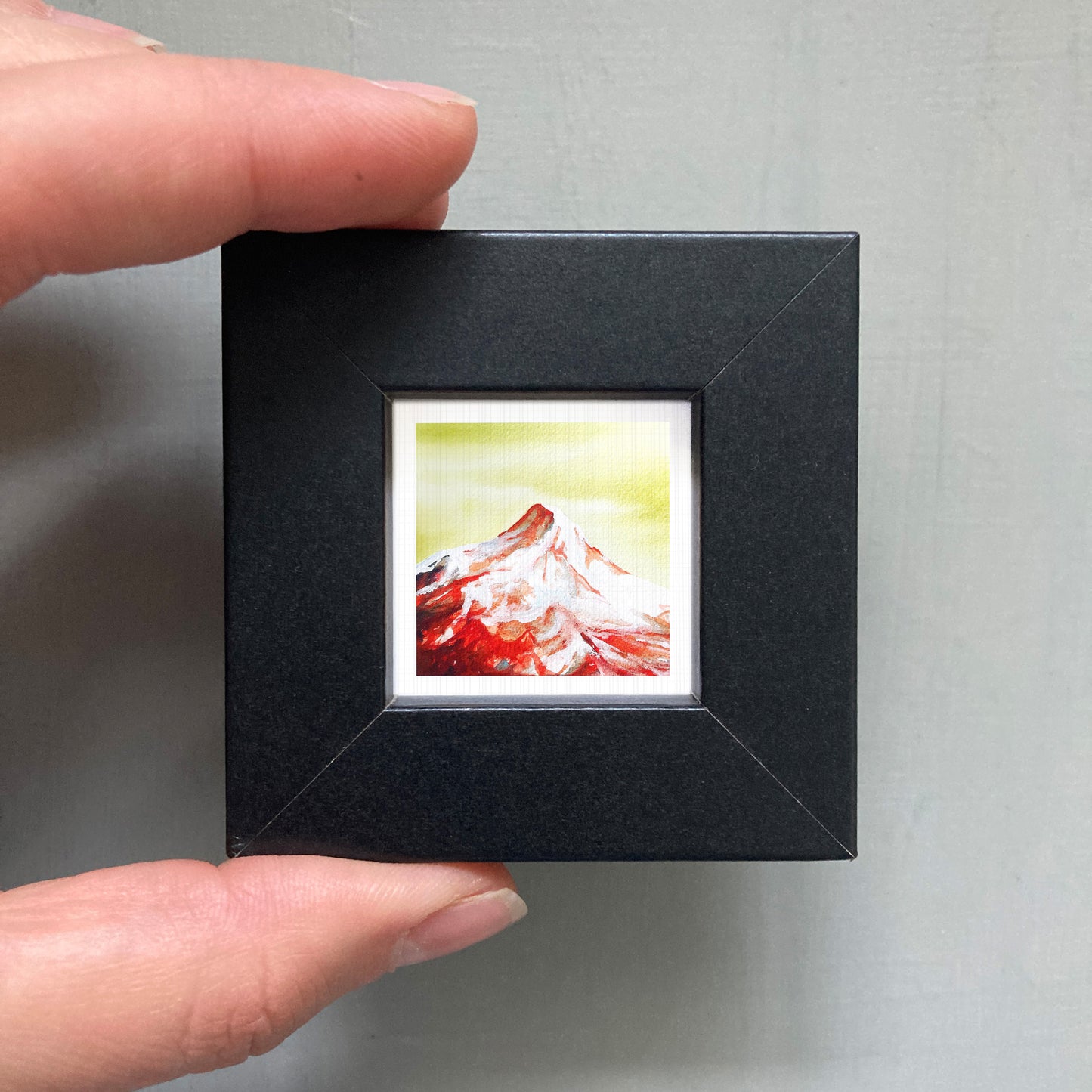 A person's hand holding a small black frame containing a framed painting of a mountain landscape.
