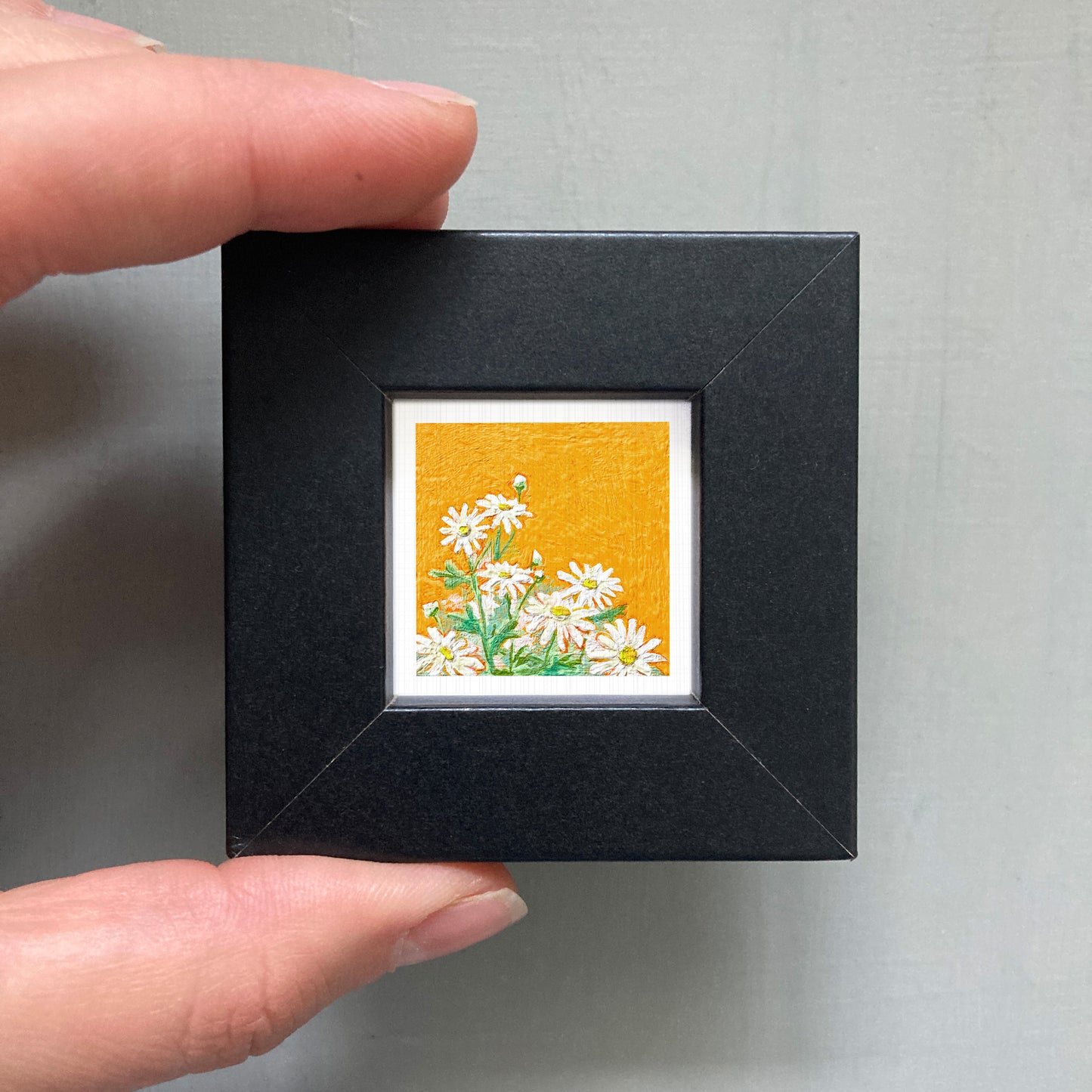 A person's hand is holding a small black frame containing a framed picture of white flowers against an orange background.