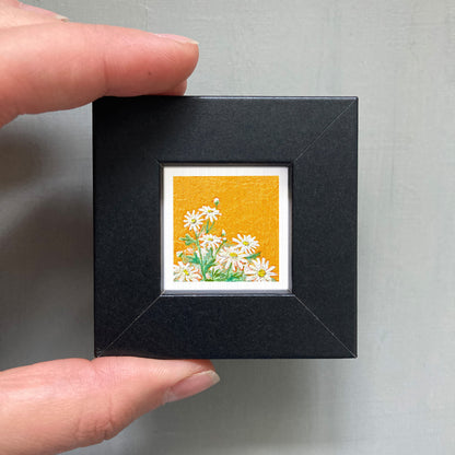 A person's hand is holding a small black frame containing a framed picture of white flowers against an orange background.
