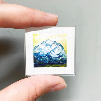 A person's hand holding a small square picture of a mountain landscape.