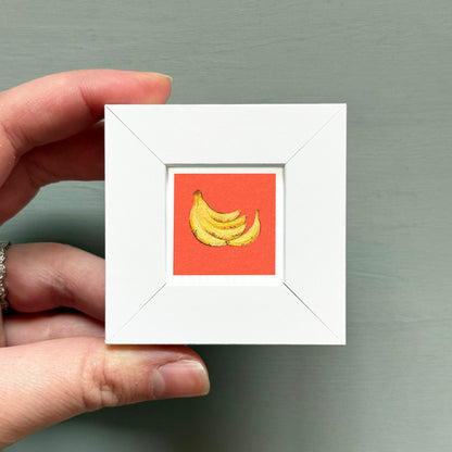 A hand is holding a small white square frame containing a vibrant illustration of a bunch of bananas.