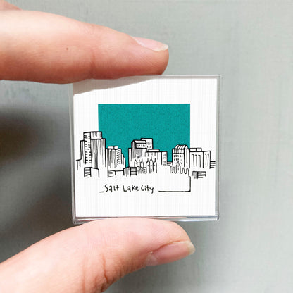 A person's hand holding a small square magnet with a drawing of a city skyline and the text "Salt Lake City" written below it.