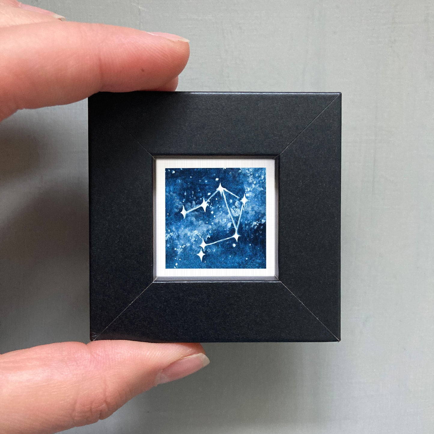 A person's hand holding a small black frame containing a framed image of a starry night sky.