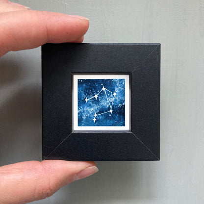 A person's hand holding a small black frame containing a framed image of a starry night sky.