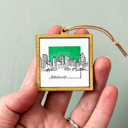 A hand holding a small wooden frame with a drawing of a city skyline and the word "Pittsburgh" written below it.