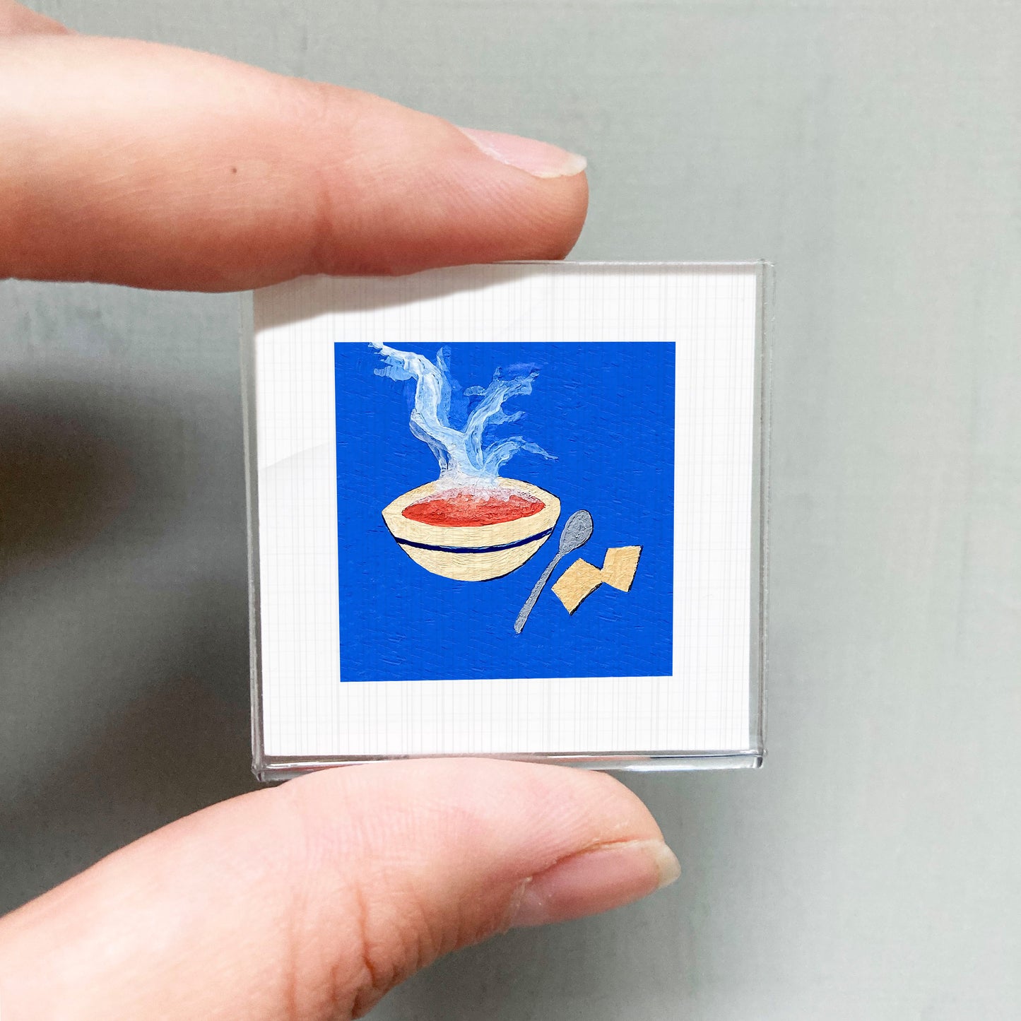 A person's hand holding a small square with an illustration of a bowl of soup and a spoon on a blue background.