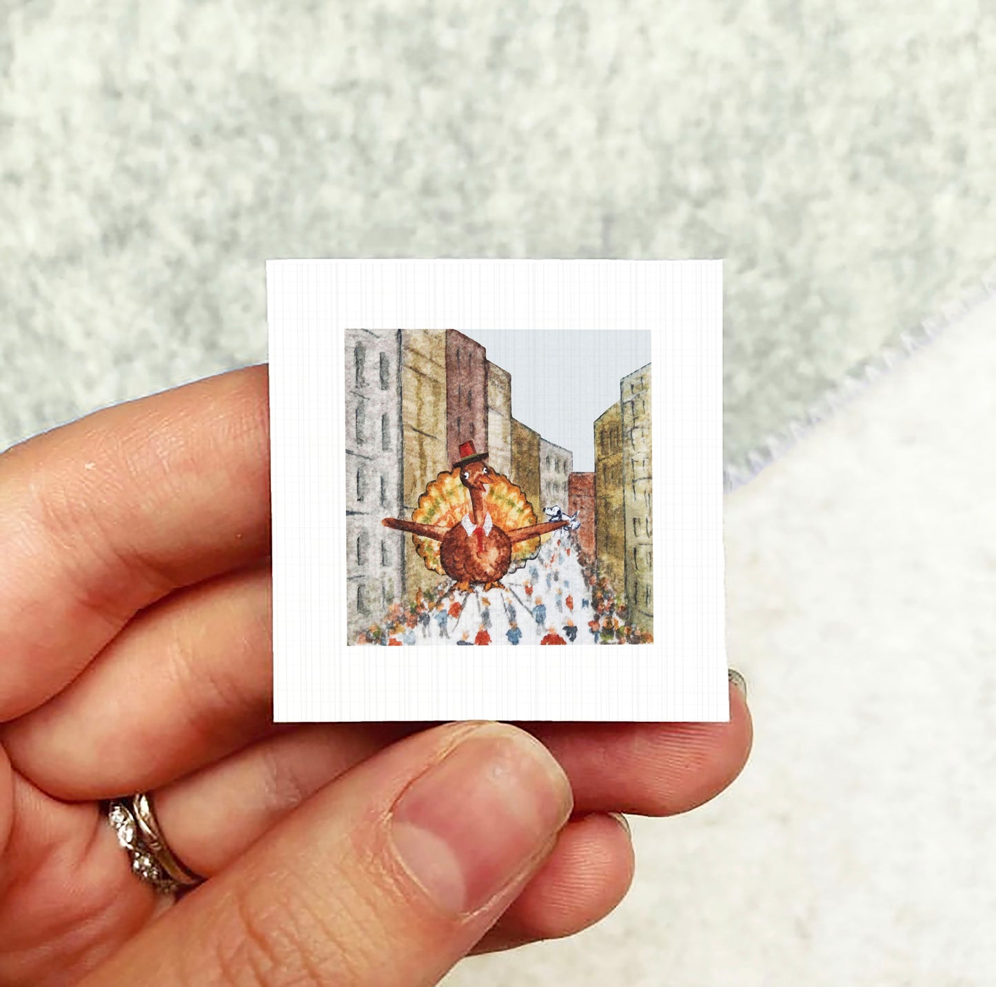 A person's hand holding a small square picture depicting a person in a colorful costume, possibly a turkey, walking down a city street.