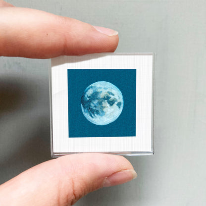 A person's hand is holding a small square with a picture of the moon on it.