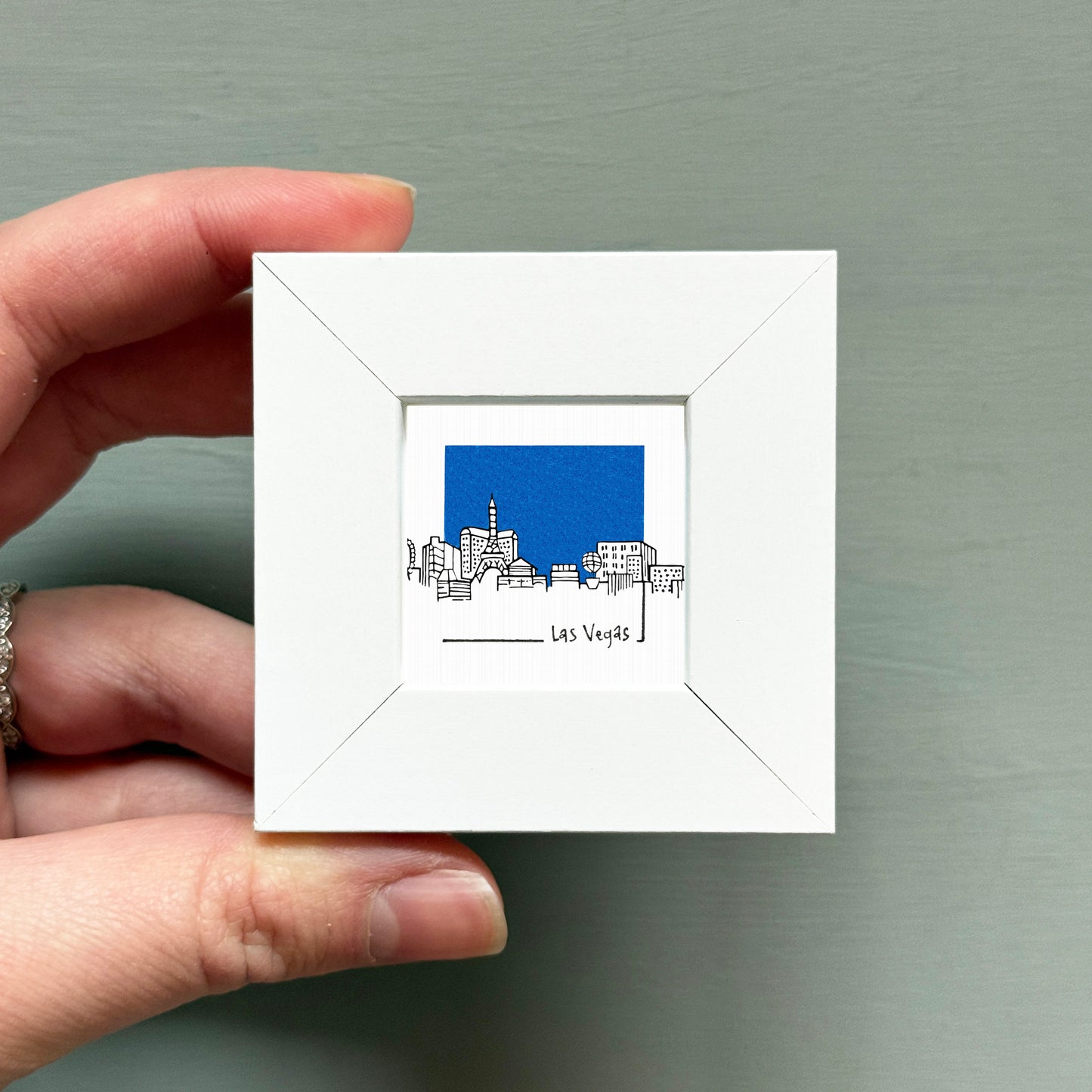 A hand is holding a small white square frame containing an illustration of a city skyline with a blue background.