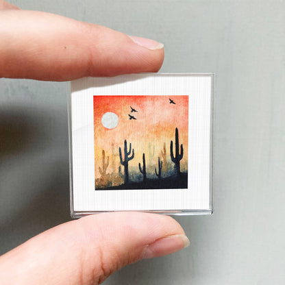 A person's hand is holding a small square picture depicting a desert landscape with cacti silhouetted against a sunset sky.