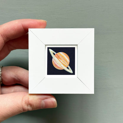A hand is holding a small white square frame containing a depiction of a planet.