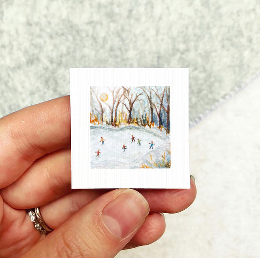 A hand holding a small square artwork depicting a snowy winter scene with people skiing on a snow-covered slope.