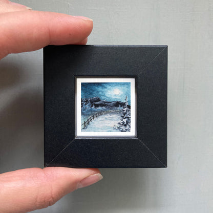A person's hand holding a small black picture frame containing a miniature landscape painting depicting a snowy mountain scene under a full moon.