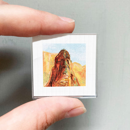 A person's hand holding a small square picture of a mountain landscape.